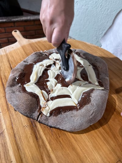 A dessert pizza, featuring a dark-colored crust topped with a swirling pattern of glossy dark brown chocolate spread and a lighter, creamy off-white substance, is being sliced on a wooden pizza peel. A person's right hand operates a pizza cutter with a black handle and a silver blade, actively pressing it into the pizza to make a cut; a previous cut is visible at the pizza's bottom edge. The scene takes place in an indoor culinary setting in Porto Alegre, Brazil, with a brick structure resembling a pizza oven visible in the left background, and a light-colored, textured wall with a white fabric element visible on the right.