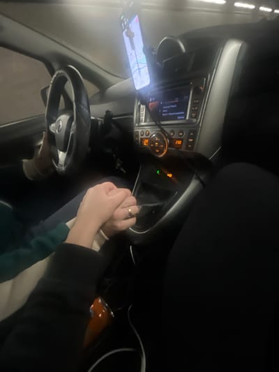 The image depicts the interior of a left-hand drive vehicle, observed from the front passenger seat perspective. The scene is dimly lit, suggesting night or an enclosed space such as a tunnel, consistent with the location of Airolo, Switzerland. A driver's left hand, wearing a dark green sleeve, rests on the steering wheel. To the right, the passenger's right hand, adorned with a gold-colored ring on the ring finger, is clasped with the driver's hand over the car's center console. A smartphone, prominently mounted on the dashboard, displays a brightly lit navigation map showing a blue route line. Below it, the car's infotainment system and climate control panel are visible, with a circular dial displaying "23.0" and a digital readout showing "230". A dark cable connects the smartphone to the vehicle's power. An orange insulated bottle is situated in a cup holder located on the center console near the clasped hands. Overhead lighting fixtures, potentially from an external environment like a tunnel, are faintly visible in the upper background.