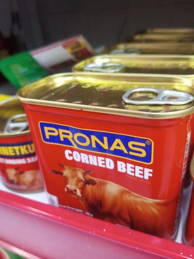 The image displays a close-up, low-angle view of multiple food cans arranged on a red shelf in a retail establishment in Naung Bo Oe, Myanmar. In the foreground, a prominent red, rectangular can of "PRONAS CORNED BEEF" is visible, featuring a blue "PRONAS" logo, white "CORNED BEEF" text, and an illustration of a light brown cow. This can has a gold-colored lid equipped with a silver pull-tab. Behind it, a linear arrangement of additional, identical "PRONAS CORNED BEEF" cans recedes into the background, becoming progressively out of focus, though their gold lids and silver pull-tabs are discernible. To the left and slightly behind the main can, a smaller, cylindrical red can labeled "CORNETKU" is partially visible. The background elements are highly blurred but suggest other packaged goods on shelves. No individuals, direct actions, or interactions are visible.