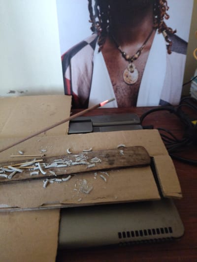 An overhead close-up shot shows a lit incense stick burning with an orange glow at its tip, angled across a jumble of cardboard. The incense stick is in the foreground, resting on top of two pieces of cardboard. One piece of cardboard, a light brown color, is positioned horizontally and appears to be a base. On top of this base, another piece of light brown cardboard is placed at an angle, also serving as a surface. Scattered across the angled cardboard are fragments of white ash from a burnt incense stick, along with a few small wooden sticks. In the background, a portion of a large poster or print is visible, depicting the upper body of a person wearing a necklace. The person's hair is styled in dreadlocks, and they are wearing a garment with a checkered pattern on the shoulders. To the right of the poster, dark power cords are visible, suggesting electronic devices are nearby. The scene appears to be set indoors, on a wooden surface, possibly a desk or table. The lighting suggests it may be daytime. The specific location within Limuru Central Ward, Kenya, cannot be determined from the visual information provided.