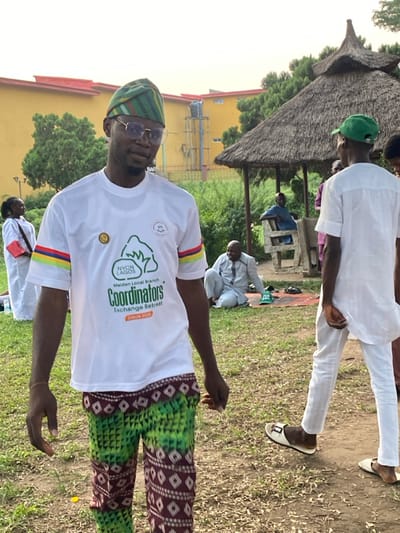 A man in a white T-shirt, green and brown pants, and a striped hat stands in a grassy area. His T-shirt has colorful stripes on the sleeves and a logo with "NYCN LAGOS Coordinators Exchange Retreat OGUN 2018". Several people are seated or standing around him. A small, thatched-roof structure is visible in the background, as is a yellow building. The man wears glasses and has a slight smile.