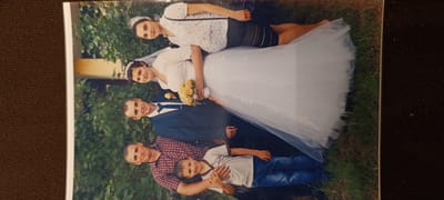 The image is a photograph of a wedding party outdoors, with greenery and grass in the background. The photograph is oriented vertically. In the foreground, a man in a plaid shirt is embracing a young boy in a t-shirt and jeans. Behind them, a man in a blue suit and tie stands next to a bride in a white wedding dress, holding a bouquet of yellow and orange flowers. A woman in a white, patterned top and dark skirt stands behind the bride. The scene appears to be a casual family gathering for a wedding celebration. The photograph is slightly faded and has a vintage quality. There is no visible indication of the location being in Winnenden, Germany.