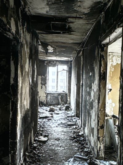 The image depicts a dark, damaged hallway within a building. The walls are charred and peeling, revealing lighter material underneath. Debris, including what appears to be broken plaster and ash, covers the floor. The ceiling is also heavily damaged and blackened. At the end of the hallway, a window is visible, letting in a dim, diffused light. The overall impression is one of severe destruction, likely caused by a fire. No people or specific objects indicating recent activity are visible. The location is identified as Neubrandenburg, Germany.