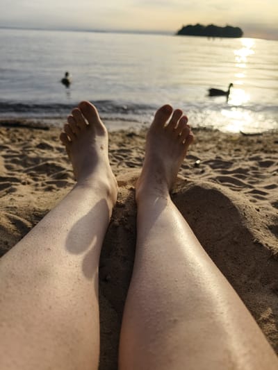 The image shows a first-person perspective of a person's legs and feet on a sandy beach. The person is lying down, with their legs stretched out towards the water. In the background, there is a body of water, likely a lake or sea, with two ducks swimming. A small island is visible in the distance, silhouetted against the setting sun. The lighting suggests it is either sunrise or sunset, with warm golden light reflecting off the water. There are no visible buildings or city structures, indicating the location is likely a natural area within or near Motala, Sweden, such as a beach along Lake Vättern.