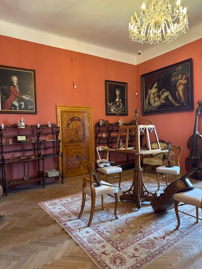 The image depicts a room in what appears to be a historical building, possibly a castle or museum, likely in Nelahozeves, Czechia. The walls are painted in a terracotta color and adorned with framed paintings of classical figures and portraits. Dominating the upper part of the room is a large, ornate chandelier. The room is filled with antique furniture including chairs with white cushions, a music stand holding a violin, a double bass, shelves displaying various objects, and an intricately designed wooden door. A patterned rug covers the wooden floor. The scene suggests a focus on music and art, with a museum setting recreating the atmosphere of a historical era.