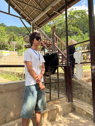 An ostrich is standing behind a metal fence, with its head raised and mouth open, near a set of stairs. To the left of the ostrich, a young male with dark, curly hair and sunglasses is standing. He is wearing a white t-shirt, blue denim shorts, and a blue lanyard with an identification tag that reads "25". His hands are clasped in front of him. To the right of the ostrich, a person wearing a black pinstripe suit and a conical hat is standing, with a white cloth draped over the fence. In the background, there is lush green foliage, trees, and a bright blue sky. The scene appears to be outdoors, possibly at a zoo or wildlife park in Định An, Vietnam, given the presence of the ostrich and the sunny, tropical environment.