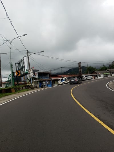 The image depicts a street scene in Bangtsho, Bhutan, under a heavily overcast sky. A dark asphalt road with a solid yellow center line curves from the mid-right to the lower left, bordered by a white line on its outer edge. Along the left side of the road, a series of single-story structures with varied facades and corrugated metal roofs are visible, appearing to be commercial or residential buildings. One storefront exhibits blue paint and text that reads "VILA SEKAMA." Multiple utility poles, laden with a dense network of overhead power lines, run parallel to the road. A large, green-framed billboard structure, featuring a yellow chevron road sign, is positioned on the far left. Several vehicles are parked along the left shoulder of the road, facing away from the viewer: a white and blue utility van, a dark gray SUV, and a silver minivan. In the background, mountains are partially obscured by low-hanging clouds. No individuals are visible within the scene.