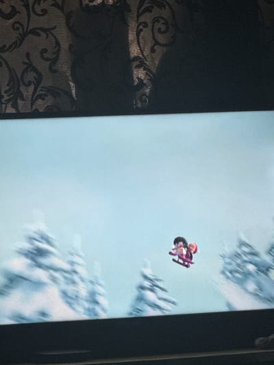 The image shows a television screen displaying a cartoon animation of two figures on skis, appearing to be in mid-air against a backdrop of snow-covered trees and a pale blue sky. The figures are small and stylized, with one wearing a pink hat and the other with dark hair. The animation has a soft, slightly blurred quality, suggesting it might be a scene from a children's movie or show. The lighting on the screen is bright, and the screen itself is situated in front of a dark, textured curtain. The overall scene is artificial and appears to be captured from a viewing distance, as the edges of the screen and a dark frame are visible. There is no indication of any real-world location in Veselinovo, Bulgaria, or any other geographical context within the image itself.