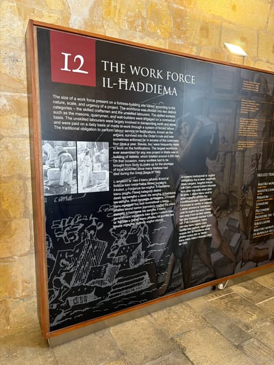 The image shows a display panel with historical information about the workforce in Valletta, Malta. The panel features text in English and Maltese, accompanied by black and white illustrations and photographs. The English text discusses the size and categories of the workforce, including skilled craftsmen and unskilled labourers, and their payment methods. It also mentions the obligation of forced labour and the importation of workers from Sicily. The Maltese text appears to be a translation or a parallel account.
The photographs depict workers engaged in construction, possibly handling large stone blocks. The illustrations show scenes of building and labor, including figures working with tools and carrying materials.
The display panel is framed in wood and is positioned against a wall constructed of rough-hewn yellow stone blocks. A bright, adjustable spotlight is mounted on the wall above the panel, illuminating the exhibit. The floor beneath the display is paved with flagstones. The overall impression is of an exhibit within a museum or historical site, detailing the monumental construction efforts undertaken in Valletta.
