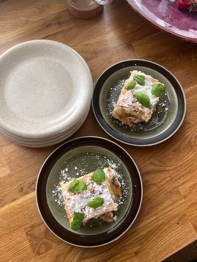 A stack of speckled, light-colored plates sits on a wooden table. To the right of the plates, two dark-rimmed, green plates each hold a slice of strudel, dusted with powdered sugar and garnished with fresh mint leaves. The strudel appears to have a flaky pastry crust and a filling of sliced fruit. The overall scene suggests a dessert being served for enjoyment. This image was taken in Hämeenlinna, Finland.