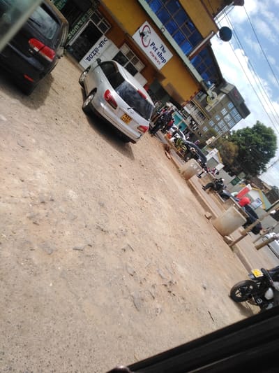 The image depicts an unpaved, dusty urban street scene in Kikuyu Township ward, Kenya, captured from a tilted, slightly downward perspective, seemingly from within a vehicle. The foreground consists of a heavily rutted and cracked dirt road.

On the far left, the rear of a dark-colored Volkswagen Golf hatchback is partially visible, parked facing away from the camera. Immediately to its right, a silver Nissan AD Van or Wingroad station wagon is parked, also facing away, displaying Kenyan license plate "KBX 118F."

Adjacent to these vehicles is a two-story building with a yellow facade on the upper level and large blue-tinted windows. A prominent sign on this building reads "The Pet shop," featuring a paw print logo and the phone number "0720 781 872." Below this, a green banner with white vertical text "M-PESA" is visible, and beneath it, a white sign with text that appears to be "VET SHOP" (partially obscured and possibly reversed/inverted).

Further down the street to the right, several individuals are visible. Some are standing, and others are seated on low concrete structures lining the street. One person is seated in a black jacket and black helmet. Another person appears to wear a red helmet. Various other parked vehicles, including motorcycles and cars, are scattered along the street. A black motorcycle with a yellow license plate is partially visible in the bottom right corner of the frame.

In the mid-ground, near the silver Nissan and the concrete structures, a brown chicken or bird is foraging on the ground. Beyond the immediate buildings, multi-story urban structures are visible, some with dark facades and numerous windows. Utility poles and overhead wires traverse the upper frame against a partially cloudy blue sky. Green tree foliage is visible on the right side, further down the street. The overall environment suggests a commercial area with ongoing activity.