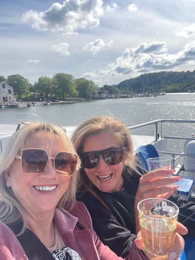 This vibrant image, captured with a sideways orientation, portrays two women enjoying a boat trip on what is likely Lake Windermere in Bowness-on-Windermere, United Kingdom. The scene unfolds on a bright, sunny day with scattered white clouds against a blue sky.

In the foreground, closer to the camera, a woman with blonde hair is smiling widely, revealing bright white teeth. She wears large, square-framed sunglasses with a gold-tone frame and is dressed in a pink velvet jacket over a lighter top. Behind her, slightly to the right, a second blonde woman, also smiling broadly, wears dark, rounder sunglasses. She holds two clear glasses, one containing a yellowish beverage, possibly white wine or a cocktail, and her fingernails are painted pink.

The women are on a boat, indicated by a metal railing visible to their right and glimpses of blue boat seating behind them. Beyond the boat, a wide expanse of sparkling water reflects the daylight. On the distant shore, a picturesque landscape features a densely forested hillside. Along the water's edge, several buildings and a marina filled with numerous small, docked boats are visible.

Both women appear happy, relaxed, and are clearly enjoying their leisure activity on the lake. The bright sunlight, calm water, and scenic backdrop contribute to an idyllic and cheerful atmosphere. There is no visible text in the image.