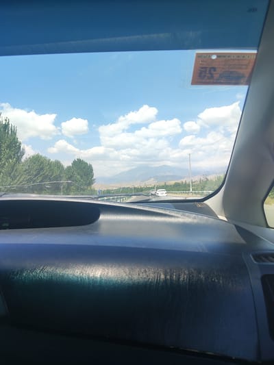 The image captures a view from inside a car looking through the windshield at a road passing through a rural landscape. The sky is blue with scattered white clouds. On the left side of the windshield, a faded orange sticker with some text and numbers is visible. The car's dashboard is in the foreground, showing a dark, glossy surface with some reflections. The road ahead is bordered by green railings and trees on either side. In the distance, mountains are visible under the bright sky. A white car is seen on the road ahead, and utility poles line the side of the road. The overall impression is of a sunny day journey through a scenic area that appears to be outside of Shahrinav, Tajikistan, given the context provided.