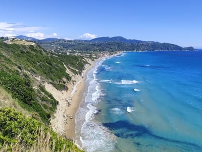 This wide-angle photograph presents a panoramic view of a beautiful coastal landscape in Agios Stefanos, Greece, under a clear, bright blue sky. A long, sandy beach forms a gentle curve along the coastline, meeting the vibrant turquoise and deep blue waters of the sea. White-capped waves break rhythmically on the shore, creating foam and movement in the water. Tiny figures of people and some small beach umbrellas are discernible on the sandy stretch.
On the left, a steep, green-covered hillside rises, transitioning into a light-colored, sandy cliff face that borders the beach. Further inland, a village or town with numerous buildings is nestled among lush green hills, extending towards hazy mountains in the distance. The sky is largely clear, with a few scattered white clouds visible towards the horizon. The scene is brightly illuminated by daylight, suggesting a sunny day. No visible text is present in the image.