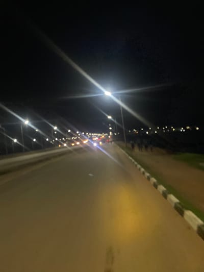 This is a nighttime, low-angle, vertical shot of a road in Gbessa, Nigeria. The road is paved and appears to be bordered by a curb with black and white stripes. To the left of the road, there is a dark area, possibly a field or unlit area, with a line of lights stretching into the distance. These lights are blurry and appear to be streetlights or lights from vehicles. There are also strong light flares emanating from some of the lights, indicating a bright light source. The overall mood is dark and somewhat mysterious due to the low light conditions and the prominent light flares. The time of day is clearly night. There is no visible text in the image.