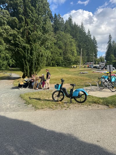 A sunny daytime outdoor scene is captured at Mosquito Creek Park in the District of North Vancouver, Canada. A group of people are gathered around a picnic table under a large, mature tree, engaged in what appears to be an art session, with easels and art supplies visible. A man in a black t-shirt and tan pants stands near the group, observing. In the foreground, a blue shared bicycle is parked, with a bike rack holding two other bicycles in the mid-ground. Beyond the park area, a street with parked cars and people walking is visible, with a densely wooded hillside and a cloudy sky forming the backdrop. The ground is a mix of gravel paths and grassy areas.