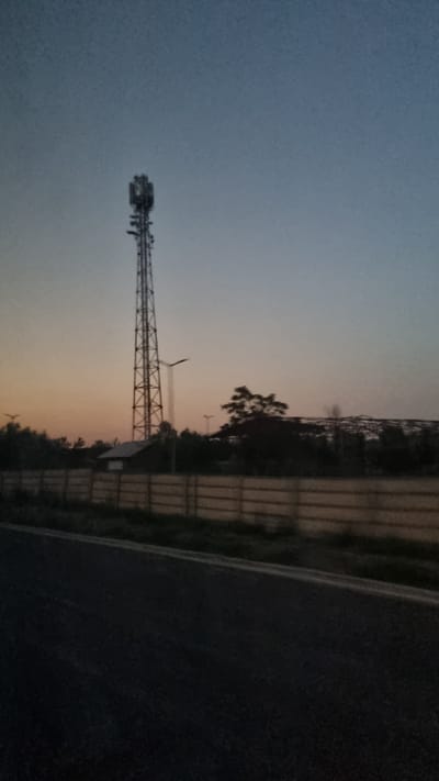 This image shows a communication tower against a twilight sky. The tower is tall and lattice-like, with antennae at the top. To the left of the tower is a small building. In the background, there are trees and a large, partially constructed structure that appears to be a building or stadium. A concrete fence runs horizontally across the middle ground. In the foreground, a dark asphalt road is visible, suggesting the image was taken from a moving vehicle. The overall impression is of an industrial or semi-developed area on the outskirts of a city, possibly at dusk.