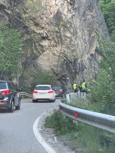 A traffic scene in Бачковски манастир, Bulgaria, shows several cars queued on a winding road next to a rocky cliff face. Two individuals wearing fluorescent yellow vests stand by a white guardrail. The registration plate "PB 9958 HX" is visible on a white car. Vegetation is abundant on the cliff face and surrounding the road.
