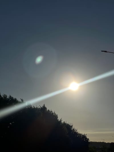 The image depicts a bright, hazy sky, likely on a sunny day, with the sun creating a strong glare and lens flare. A silhouette of trees with a dense canopy forms the right side of the frame, obscuring some of the background. In the upper left corner, a portion of a dark, pole-like object is visible, possibly a lamp post or part of a structure. A thin, distant horizontal band of lighter sky is discernible at the top of the frame, suggesting the horizon. The overall scene appears to be an outdoor setting, possibly a park or a natural area, given the presence of trees. The time of day is likely late morning or early afternoon, judging by the position and intensity of the sun. No people are visible in the frame. No text is present. The lighting conditions create a sense of diffused sunlight and a slightly muted color palette.