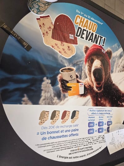 The image displays a circular promotional advertisement, approximately 30-40 cm in diameter, placed on a dark, flat surface, likely a countertop within a commercial establishment in France. The advertisement features a winter-themed graphic with a digitally rendered brown bear wearing a red knitted beanie, holding a white MaxiCoffee branded cup of steaming coffee. The bear is set against a blurred snowy mountain backdrop with conifer trees. Text in French reads "Du 1er au 31 décembre*", "CHAUD DEVANT!" (Warm Up Front!), and promotes an offer: "Dès 20€ de recharge café = Un bonnet et une paire de chaussettes offerts" (From €20 coffee top-up = A free beanie and a pair of socks). A red knitted beanie and a pair of light beige patterned socks are depicted above the bear. A bonus offer table details rewards for coffee recharges on a TotalEnergies branded card, showing "20€ + 4€ = 24€" and "30€ + 6€ = 36€". Below this, four pairs of patterned socks in various colors (light beige, green, red, dark gray) are illustrated. Fine print in French states the offer is valid at participating TotalEnergies stations in metropolitan France. The slogan "L'énergie est notre avenir, économisons-la!" (Energy is our future, let's save it!) appears at the bottom. To the upper left of the advertisement, a portion of a patterned paper cup is visible. To the left, a black rectangular sachet labeled "PAUL" rests on the dark surface. A crumpled white paper receipt partially overlaps the lower right section of the advertisement. The image itself does not provide the specific city of Urvillers, France, but indicates a TotalEnergies promotion occurring within metropolitan France.