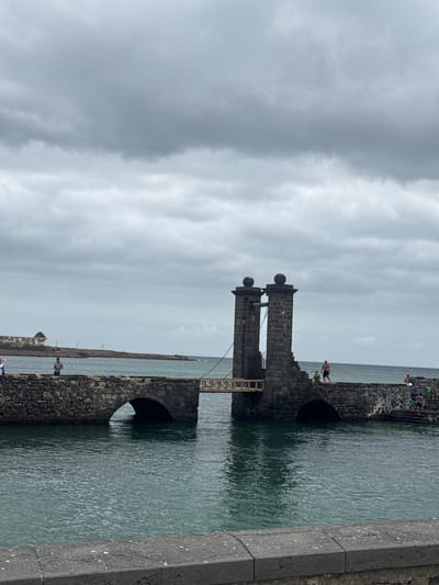 The image depicts the Castillo de San Gabriel in Arrecife, Spain. The fort is constructed of dark stone and features distinctive cylindrical turrets topped with spherical decorations. A wooden bridge connects the turrets. The fort is surrounded by water, with arched openings visible in the stone structure. Several people are present on the fort's walls, and the sky is overcast.