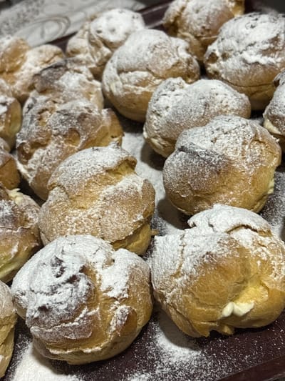 A close-up, overhead view of a tray filled with freshly baked cream puffs. The puffs are golden brown with a slightly crispy exterior and are generously dusted with powdered sugar, creating a soft, snowy appearance. Some puffs are partially obscured by others, creating a layered and abundant presentation. A few cream puffs show hints of a creamy filling peeking out from small cracks. The background is dark, likely a tray or surface, with scattered specks of powdered sugar indicating their recent preparation. The lighting is warm and natural, suggesting the pastries might be indoors, possibly in a kitchen or bakery setting in Sheerteba, Georgia. The overall impression is one of deliciousness and homemade goodness, with no people, text, or specific time of day or weather cues visible.