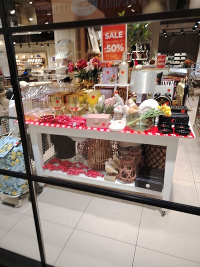 This is an eye-level, medium shot of a store window display. The display features a white table with various decorative items. On the tabletop, to the left, are several red lobster-shaped decorations. Next to these are pink boxes, and further to the right, a vase with yellow flowers, a stack of black bowls, and a quilted black bag. Behind the table, an assortment of merchandise is visible, including lamps, vases, and other decorative items.
A large red sign on the left side of the display reads "SALE -50%". A smaller red sign on the right also says "SALE 50%". The store appears to be a home decor or gift shop. The window is framed by thick black bars, suggesting it's a storefront. The background shows other shelves of products, indicating this is a retail environment. The floor tiles are visible in the foreground, and the overall lighting suggests it is daytime. The display is intended to attract customers by highlighting sale items and aesthetically pleasing products. No people are visible within the frame.
