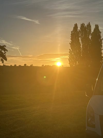A low-angle, portrait-oriented photograph captures a sunset over a field in Tewkesbury, United Kingdom. The sun, a bright orb of light, is positioned in the upper center of the frame, casting a warm, golden glow across the landscape. Wispy clouds streak across the pale blue sky, with contrails from aircraft visible. In the mid-ground, a line of silhouetted trees stands against the horizon, with a few more scattered trees in the distance. The foreground consists of a grassy field, bathed in the sunset's light, with a portion of a white vehicle visible on the right side of the frame. The overall impression is one of a serene and tranquil evening.
