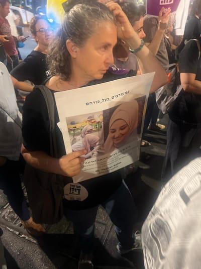 An outdoor night scene in West Jerusalem, Israel, depicts a crowd during a public gathering. In the foreground, a middle-aged woman with dark, graying, curly hair, pulled back, holds a white protest sign. She wears a dark short-sleeved top, dark pants, and a dark shoulder bag. Her right hand is raised to her head, and her left hand supports the sign. The sign features two images of a young woman wearing a hijab: a left image showing her in a medical or laboratory setting, and a right image as a smiling portrait. Text on the sign includes Hebrew characters, with visible phrases such as '#מעורבים_בכל_כוחם' (Involved with all their might) and 'Dr. Thabet Salim' in both Hebrew and English. A circular sticker on the woman's shirt displays the Hebrew phrase 'מתי נביא די?!' (When will we say enough?!). The background contains a blurred gathering of other individuals, artificial lighting, and a reflective ground surface, possibly wet asphalt or cobblestones. Partially visible urban elements include a light-colored vehicle and a purple banner in the distance.