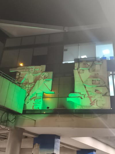 Here is a detailed description of the image:
The image shows a building interior in Guatemala City, Guatemala. The architecture is modern, with gray, white, and blue hues in the lower sections. Large panels with stylized, low-relief artwork are illuminated with bright green light. The artwork appears to be of Mayan or other indigenous Guatemalan origin, possibly replicas of historical carvings. Behind the panels are windows into what appears to be an office space, with lights on and blurry shapes of people visible inside.