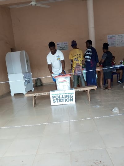 An indoor scene within a polling station in Ajalli, Nigeria, shows several individuals participating in or observing an election. The room features light beige/peach walls and a tiled floor. A white ceiling fan is visible overhead.
In the left foreground, a young man wearing a white t-shirt with "REAL LIFE" text and blue shorts is positioned over a clear plastic ballot box with a red lid, appearing to cast a vote. To his left stands a two-part white voting booth with a green logo, and a green backpack rests on the floor nearby.
A white rope or string acts as a barrier across the foreground. Behind this barrier, a wooden bench displays "LEGION OF M..." carved into it, and in front of the bench, a sign reads "INEC INDEPENDENT NATIONAL ELECTORAL COMMISSION POLLING STATION" along with "INEC POLLING UNIT 00 A UMWUKWU."
Further back, to the right of the man casting his vote, another man in a yellow t-shirt with a printed image and a purple beanie stands looking forward. Next to him, a woman in a dark blue patterned dress looks towards the foreground. To her right, another woman in a black and white patterned top stands near a wooden table.
On the background wall, multiple white posters related to the Independent National Electoral Commission (INEC) are affixed, including one prominently displaying "VOTING IN PROGRESS." Debris, including a plastic bag, is visible on the tiled floor in the lower right.