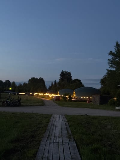 The image depicts an outdoor scene at dusk, possibly in the city of Zamosh'e (Zaklinskoe S/P), Russia. A wooden boardwalk leads towards the viewer from the foreground, bordered by grass. In the midground, a gravel path curves through an area illuminated by string lights. To the left of the path, bicycles are parked under a simple canopy. Further down the path, several domed structures with lights are visible, suggesting a glamping or event site. A red car is parked near the domed structures. Trees and foliage surround the area, indicating a semi-rural or park-like setting. The sky is a dusky blue.