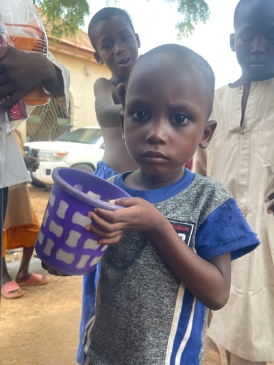 The image depicts a close-up of a young Black child in the city of Waziri A, Nigeria, holding a purple and white patterned bucket. The child is wearing a gray and blue t-shirt with white stripes on the side. The child's expression is serious, with dark eyes looking directly at the camera. In the background, other children and adults are partially visible. To the left, an individual is holding a clear plastic bag containing items. Further in the background, a white SUV is parked, and part of a building with an arched window is visible. The ground appears to be dirt. The overall scene suggests an outdoor setting in a community.