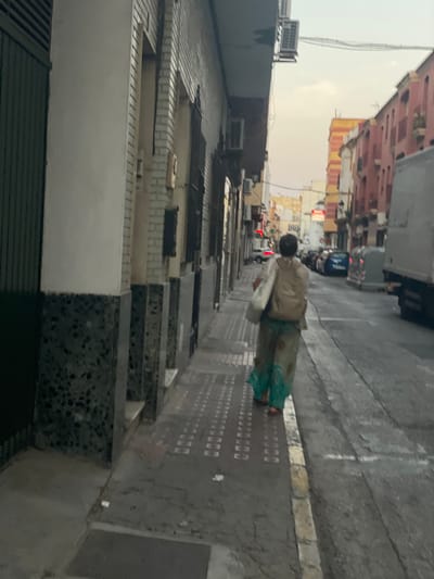 The image depicts a street scene in La Línea De La Concepción, Spain, during what appears to be late afternoon or early evening. A person, viewed from behind, walks away from the camera down a paved sidewalk. The individual is wearing a light-colored, possibly beige, backpack and a long, flowy garment that appears to be a dress or skirt with teal accents. The sidewalk is lined with textured paving stones. To the left of the sidewalk is a building with a white facade, dark green metal doors, and a lower section clad in dark speckled material. The street itself is asphalt and has parked cars and a large white truck visible in the distance. The buildings lining the street are multi-story and exhibit architectural styles typical of Spanish cities, with some facades painted in vibrant colors like pink and orange. The sky is overcast, suggesting a diffused light source.
