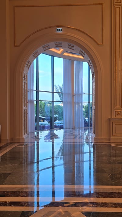 The image shows a grand arched doorway with ornate gold trim, leading to a large window. Through the window, a bright blue sky with scattered clouds is visible, along with lush green trees and foliage. White sheer curtains hang on either side of the window, softening the natural light. The floor is highly polished marble, reflecting the scene outside and the architectural details of the interior. There is an illuminated exit sign above the archway. The overall impression is one of luxurious and sophisticated interior design, likely in a hotel, resort, or similar establishment. The location is identified as Agios Epiktitos, Northern Cyprus / Cyprus. There are no people visible in the image.