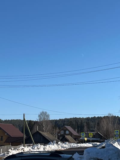 The image displays an outdoor, daytime scene within the city of Сундур, Russia, under a clear blue sky. A distinct crescent moon is visible in the upper right quadrant of the sky. Multiple dark, parallel power lines traverse the sky horizontally across the upper and middle sections of the frame. The foreground contains accumulations of dirty snow, partially obscuring the top portion of a dark-colored vehicle, indicating the perspective is from inside a vehicle.
In the middle ground, a cluster of wooden residential buildings with varying roof designs (some brown-shingled, others dark and weathered) are present. A light green utility pole stands prominently on the left side. Behind the structures, bare deciduous trees are visible, intermingled with a dense coniferous forest that forms the distant background, spanning a gentle incline. Several yellow, diamond-shaped traffic signs are present along what appears to be a street or road; these include signs indicating pedestrian crossings (featuring a walking figure) and a sharp left curve. No individuals, explicit actions, or interactions are observable. The presence of snow and bare trees suggests a late winter or early spring season.