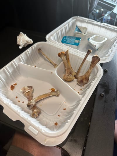 A partially consumed meal, consisting of chicken wing bones and small condiment containers, rests in a white plastic takeout container. The container is divided into sections, with some sections empty and others holding the remnants of food. A crumpled white napkin lies to the left of the container. The scene is set against a dark, textured surface, possibly a table. The lighting is dim and appears to be artificial. There is no discernible indication of a specific location within Raphine, United States; this information is not present in the visual data.