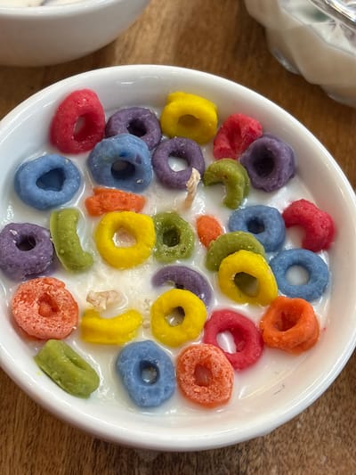 A white bowl filled with colorful cereal loops and milk sits on a wooden surface. The cereal pieces are red, orange, yellow, green, blue, and purple. The scene appears to be indoors. There is no indication of the location being in Chorrillos, Peru.