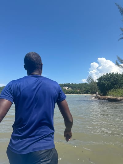 An image captured in Rio Bueno, Jamaica, depicts an adult male from a rear perspective, standing in shallow, murky water. The subject has dark skin, short dark hair with visible grey, and is wearing a royal blue short-sleeved t-shirt and dark shorts. The water surface shows ripples and scattered minor debris. In the mid-ground, a body of water extends towards a distant shoreline. The immediate right edge of the frame features a rocky bank with dense green foliage. To the left and in the background, a sandy beach is visible, backed by verdant, tree-covered hills. Several light-colored structures are discernible on the distant hillsides. The sky is clear blue, with a prominent white cumulus cloud formation positioned in the upper right. The tips of evergreen tree fronds are visible at the extreme upper right corner. The scene is illuminated by direct sunlight.