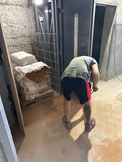 In Djerman, Bulgaria, a person wearing a green t-shirt and black and red shorts is sweeping the floor of a basement or utility area with a long-handled broom. The floor is concrete and has a light brown, dusty appearance with white powdery residue in patches. To the left, a stack of several large white sacks, some torn and revealing a brown powdery substance, are placed on a wooden pallet. Behind the sacks, a metal cage or barrier is visible. In the background, a large, dark metal structure, possibly an elevator shaft or industrial machinery, is present. The walls are rough stone and concrete. Lighting appears to be from overhead fixtures.