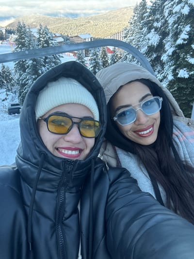 The image displays two females taking a selfie while seated on a chairlift on Uludağ mountain, Bursa, Turkey. The individual on the left wears a white knit beanie, a black hooded puffer jacket, and yellow-tinted sunglasses with dark frames, smiling with red lipstick. The individual on the right wears a light tan hooded puffer jacket, a light-colored patterned scarf, and blue-tinted clear-framed sunglasses, also smiling with red lipstick. The background reveals a snowy mountain landscape, including numerous snow-covered coniferous trees, several multi-story resort buildings, and distant snow-capped peaks under a partly cloudy sky. A metallic chairlift bar is visible horizontally in the upper background.