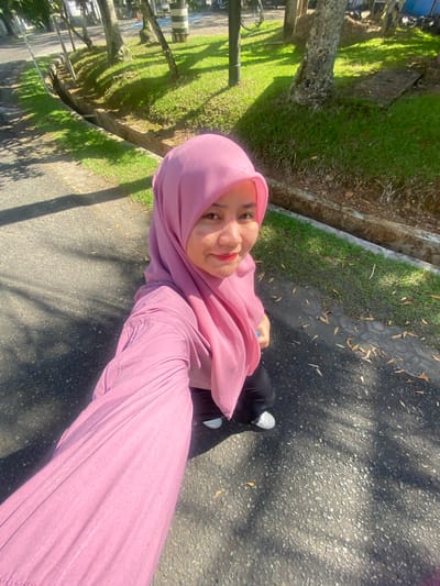 A young woman wearing a pink hijab and a pink long-sleeved shirt is captured in a high-angle selfie outdoors. She is standing on an asphalt path, possibly in a park or recreational area in Banjarbaru, Indonesia, given the presence of trees, grass, and a drainage ditch. Sunlight casts shadows on the ground, indicating it is daytime. The woman's face is partially visible, and she appears to be smiling. She is wearing black pants and white sneakers. The background features a well-maintained grassy area with scattered leaves, and several trees. The overall impression is a casual, sunny day.
