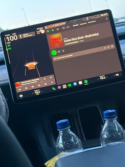 The image shows the interior of a car, likely a Tesla, with its large central touchscreen display prominently in focus. The screen displays navigation information, music playback, and car controls. The car is in motion, indicated by the navigation view showing a car icon on a road. The speed displayed is 100 km/h. The time shown on the screen is 21:46 and the temperature is 17°C. Music is playing, identified as "Judas Kiss (feat. SkyDxddy)". Two plastic water bottles with blue caps are visible in the center console. The overall environment suggests a daytime scene, possibly overcast, given the lighting. The location is identified as Sponvika, Norway.