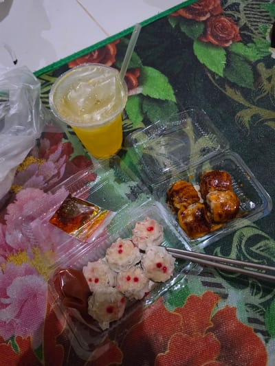 The image shows a takeaway meal, likely for one person, set on a table covered with a floral-patterned tablecloth. The meal consists of several items in clear plastic containers. In the upper right is a container with three pieces of what appear to be spring rolls or some kind of pastry, glazed with a dark, reddish-brown sauce. To the right of this is another container holding several small, steamed dumplings, possibly siomai, each topped with a small red dot. There is also a small plastic bag containing a chili oil sauce. In the lower left, a yellow cup with a straw holds a light-colored beverage, possibly juice or iced tea, with ice cubes visible. A white plastic bag lies beneath the cup and containers. The background features a dark green tablecloth with a bold floral pattern, featuring red roses on the left and large orange and pink flowers on the right. The lighting suggests it is indoors, possibly in the evening. The setting appears casual and domestic, consistent with a takeaway meal enjoyed at home in Pintomakmur, Indonesia. There are no people visible. The overall impression is one of a simple, convenient meal.