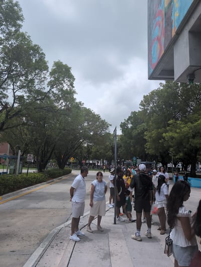 A group of people, some standing and some in a line, are gathered outdoors under a cloudy sky. The setting appears to be a street or sidewalk area with trees lining the background. To the left, there's a bus stop with signage. In the distance, a mural is visible on a building. The people are dressed in casual attire, with a mix of ages and genders represented. Some individuals are looking at their phones, while others are engaged in conversation. The overall atmosphere suggests a public gathering or waiting area. The location is identified as Gladeview, United States.