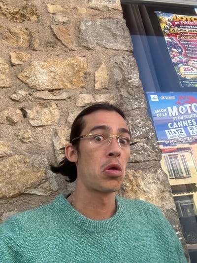 The image shows a person with dark hair, wearing glasses and a green sweater, standing in front of a stone wall. Behind the wall, there is a window with posters visible. The posters advertise "LOTO DE PAQUES DU ROTARY" for April 4, 2026, in Cannes, France, mentioning prizes like scooters and TVs. Another poster advertises "SALON DE LA MOTO ET DU SCOOTER CANNES" for April 11 & 12, 2026, at Stade Coubertin, noting the presence of the "MOTO CLUB DE CANNES." The setting appears to be in Mougins, France.