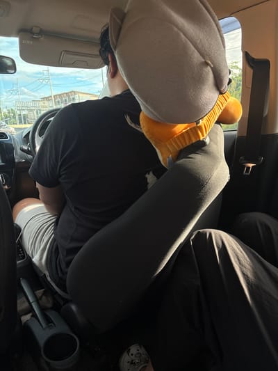The image shows the interior of a car, viewed from the back seat. A person is driving, wearing a black t-shirt and grey shorts. Their legs are crossed, with one leg extending towards the camera. A stuffed animal is placed on the passenger seat. Outside the car, power lines and buildings are visible, suggesting an urban or semi-urban setting. The location is identified as Tha Sala, Thailand. The overall lighting indicates it is daytime.