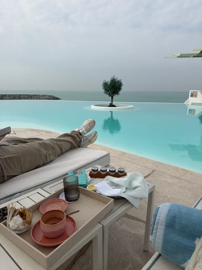The image depicts an outdoor waterfront scene in Ghadeer Al Tayr, United Arab Emirates, characterized by an infinity pool overlooking the sea under an overcast sky.
In the foreground, the lower half of an individual's body is visible, clad in light tan cargo pants and light-colored athletic shoes, with legs crossed at the ankles, resting on a light grey cushioned lounge chair with light blue piping. Adjacent to the lounge chair, a light-colored rectangular side table holds a beige serving tray containing an empty pink ceramic cup and saucer, an additional pink saucer, and a tall, ribbed dark grey drinking glass. A clear glass bowl with individually wrapped light brown items and crumpled white paper rests beside the tray.
On the table to the right, a wooden caddy with four small glass jars is present. Next to it, a white plate with food remnants is partially covered by a light-colored napkin, and a small dish containing a yellow substance is visible. A fork and knife are wrapped in a light teal napkin. To the far right, a light blue and white striped towel is folded on another lounge chair, partially visible.
The midground is dominated by a clear, turquoise infinity pool. A small, circular white platform within the pool features a small, green-leafed tree, its reflection visible in the water. Beyond the pool, the calm, muted greenish-blue sea extends to the horizon, meeting an overcast, light grey sky. A man-made rock breakwater extends into the sea on the left side. On the far right, a white structure with tiered, angular awnings is partially visible, alongside a white panel displaying blue iconography.