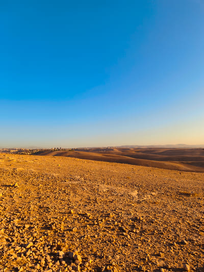 The image depicts a wide, arid landscape under a clear blue sky. The foreground is dominated by a rough, rocky ground surface, illuminated by warm, golden light, suggesting either sunrise or sunset. In the middle ground, gently rolling hills of a similar arid terrain extend towards the horizon. Distantly, a settlement or city is visible, characterized by buildings and structures spread across the landscape, appearing somewhat hazy against the muted tones of the hills. The sky is a gradient of bright blue at the top, transitioning to a lighter, warmer hue closer to the horizon, indicating the presence of the sun. The overall scene is one of a dry, sparsely vegetated environment with a human settlement in the distance. The location is identified as the city of Keidar, Palestinian Territories.