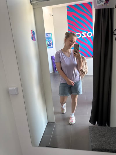A young woman is taking a mirror selfie in a fitting room, located in the city of Vyazniki, Russia. She is wearing a light purple v-neck t-shirt, denim shorts, white socks, and pink Crocs. She is holding a smartphone in her right hand and a light brown backpack is slung over her left shoulder. The fitting room has white walls and grey tiled flooring. In the background, there is a large poster with a pink and blue wavy pattern and the word "OSO" in white letters. A dark grey curtain hangs to the right of the woman, and a grey upholstered bench is visible in the bottom right corner.